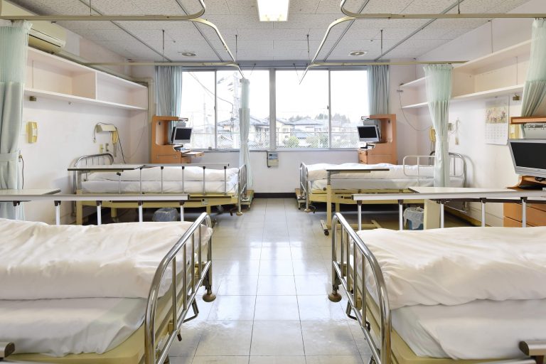 介護施設の多床室。複数のベッドが整列し、窓からは自然光が入る明るい病室環境。